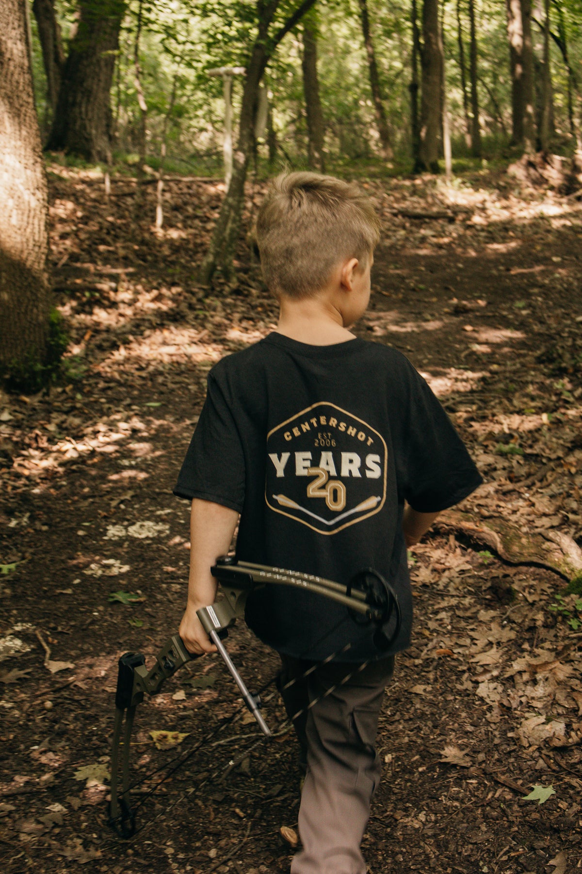 A young boy seen from behind walking on a wooded forest path, wearing a black t-shirt with a gold and white 'Centershot 20 Years' graphic on the back, holding a bow.
