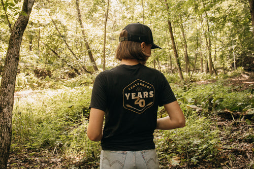 A person standing in a sunlit forest, viewed from behind, wearing a black t-shirt with a large beige and gold graphic on the back that reads Centershot 20 Years.