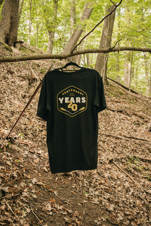 A black short-sleeve t-shirt hanging on a hanger from a tree branch in a wooded forest setting. The shirt features a gold and white graphic on the front that reads 'CENTERSHOT 20 YEARS'.