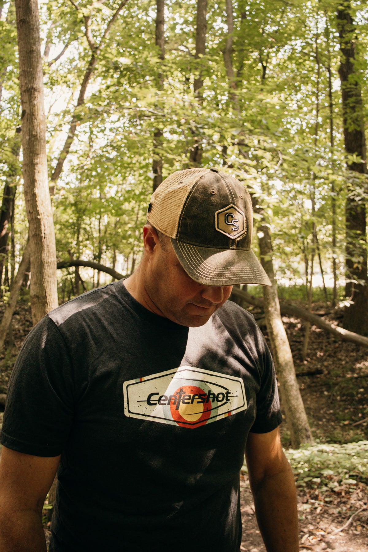 A man wearing a tan and green trucker hat and a black t-shirt with a 'Centershot' logo, standing in a wooded forest setting.