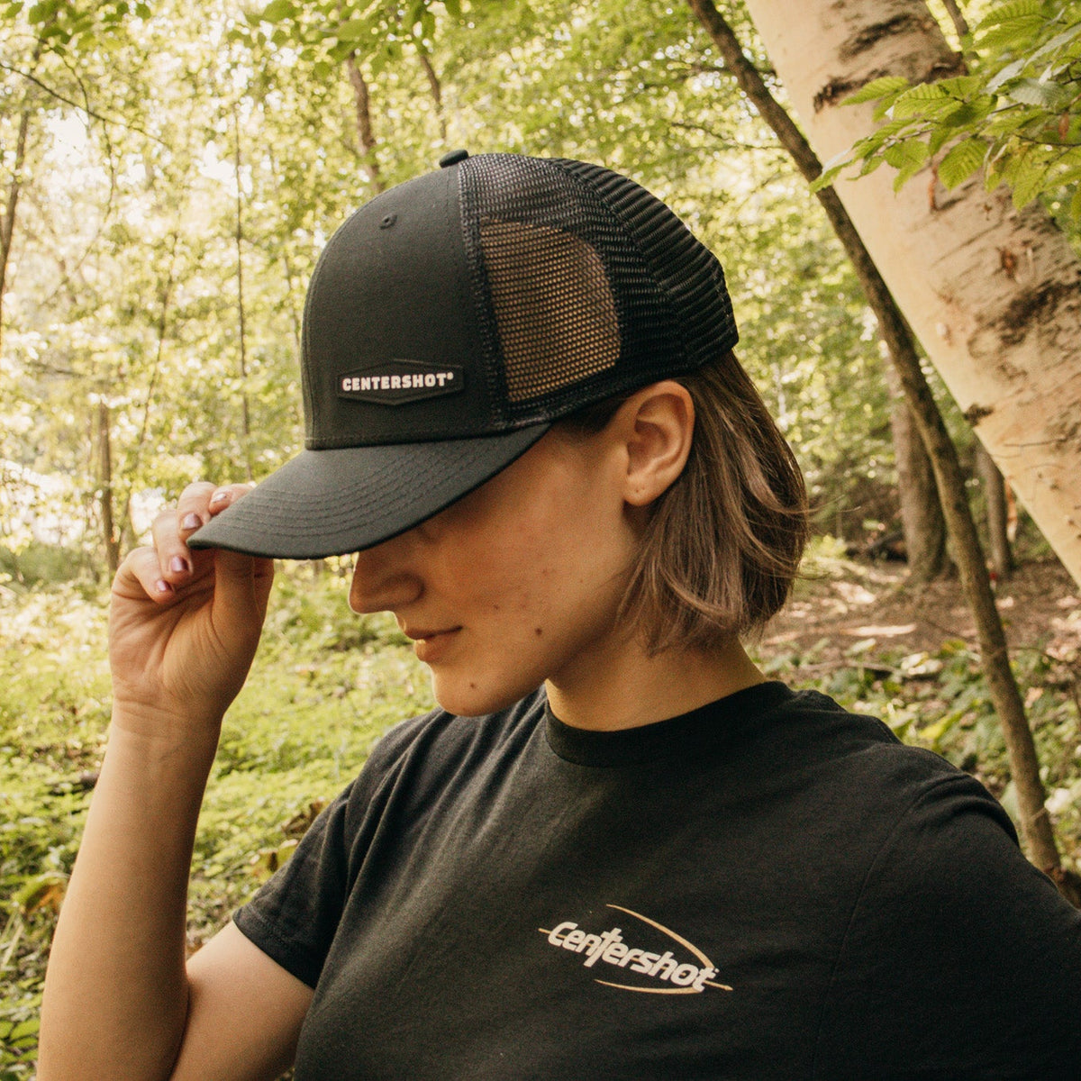 A woman wearing a black trucker hat and a black t-shirt with a white 'Centershot' logo, standing in a sunlit forest with birch trees.