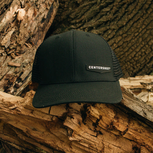 A black trucker hat with a black mesh back and a small black patch on the front that reads 'CENTERSHOT' in white, resting on weathered tree logs outdoors.
