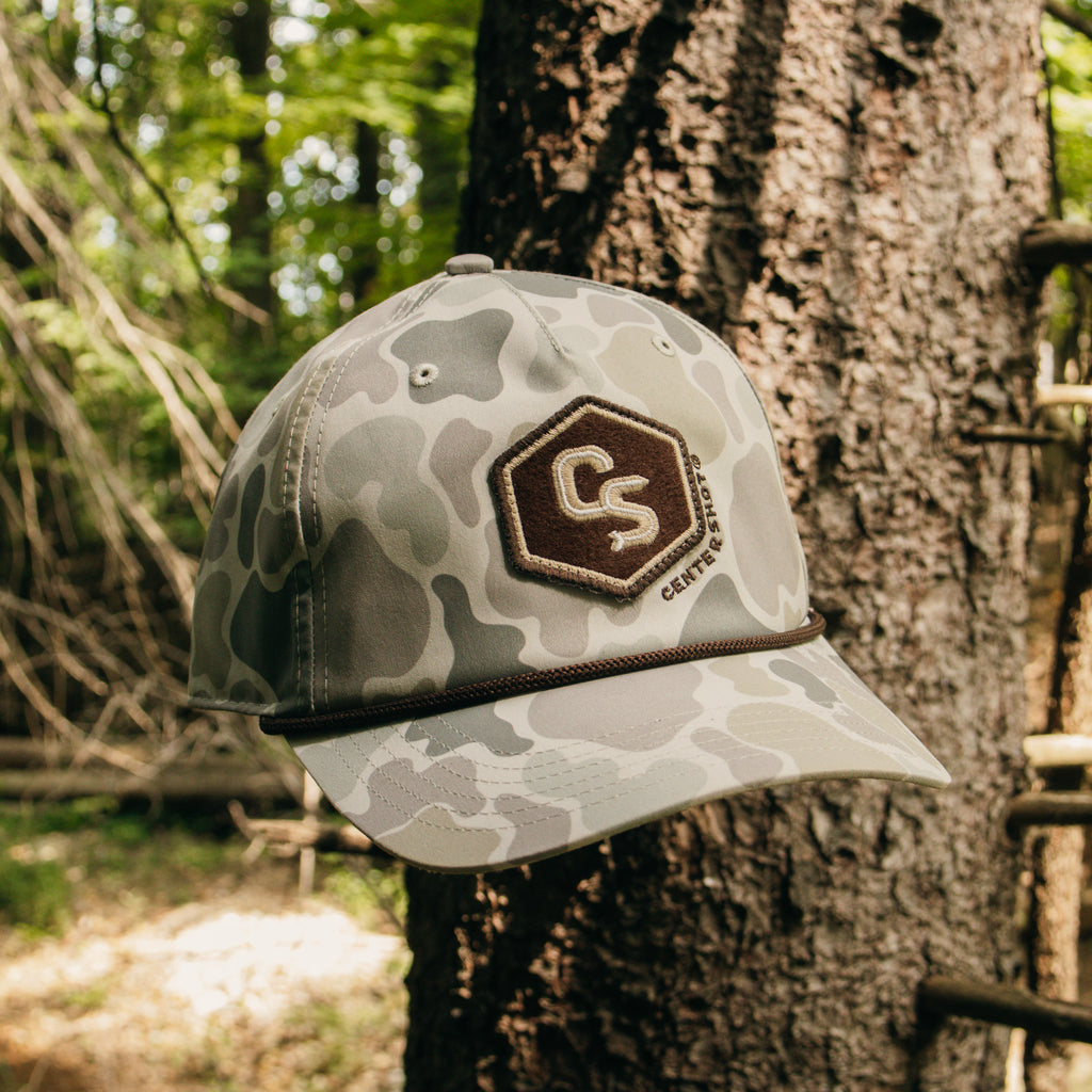 A camouflage baseball cap with a brown embroidered patch featuring the letters 'CS' and the brand name 'Centershot' hanging from a tree outdoors.