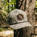 A camouflage baseball cap with a brown embroidered patch featuring the letters 'CS' and the brand name 'Centershot' hanging from a tree outdoors.