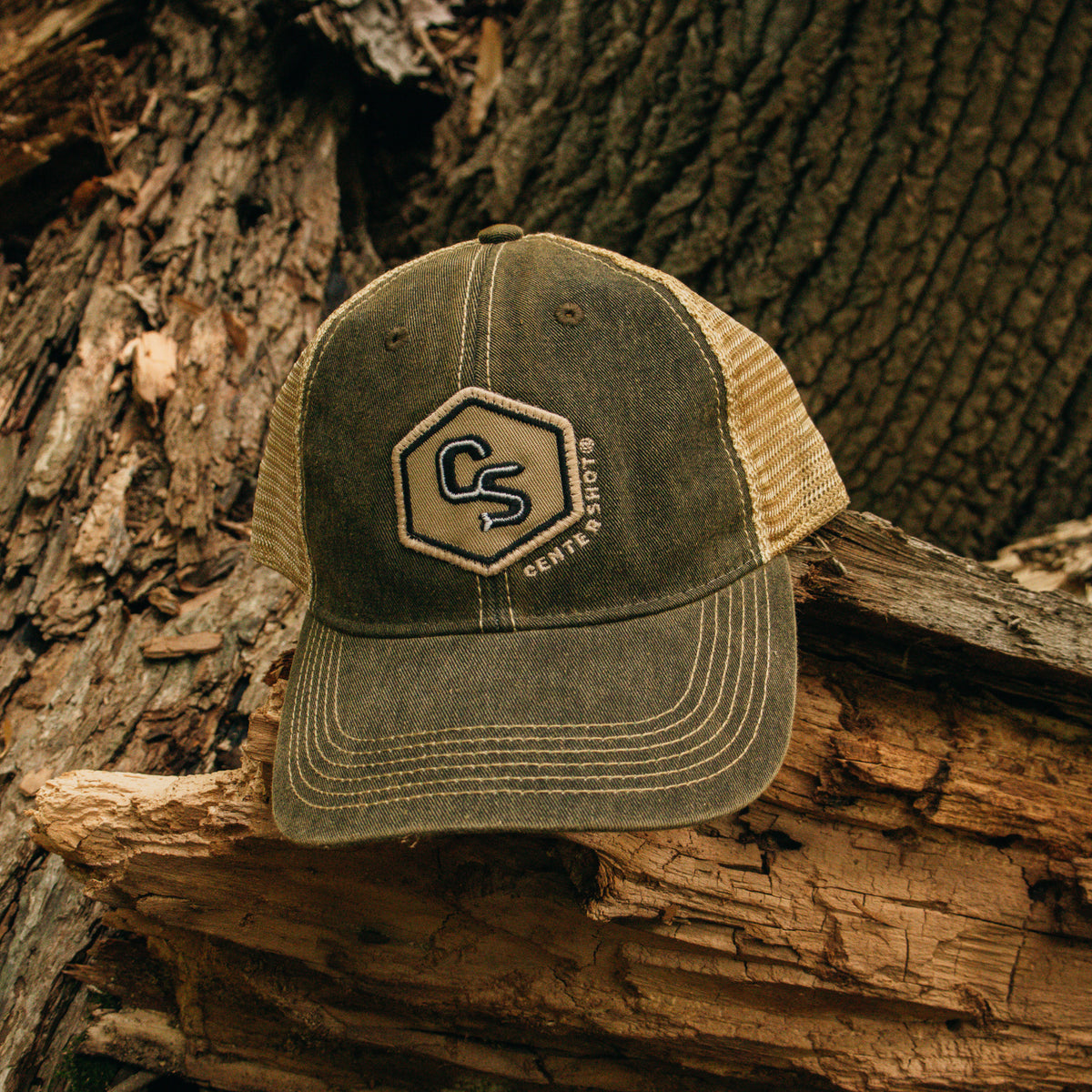 A trucker-style baseball cap with an olive green front panel and beige mesh back, resting on a piece of weathered wood against a tree trunk background. The front features a hexagonal patch with the letters 'CS' and the text 'Centershot'.