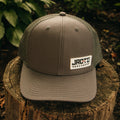 A gray trucker hat with a white 'JROTC ARCHERY' logo patch on the front, displayed on a rustic wooden tree stump with a lush green forest background.