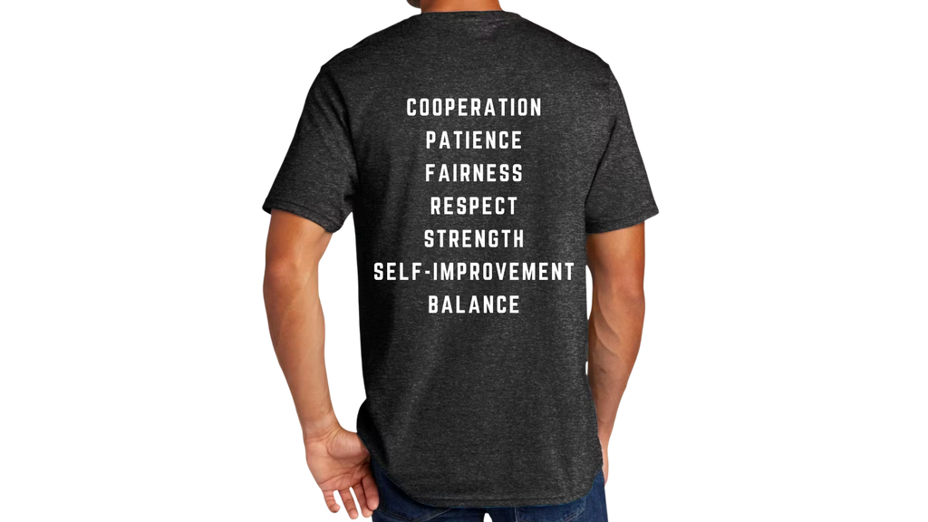 A man wearing a dark heather gray t-shirt, viewed from the back. The shirt features a vertical list of motivational words in white block lettering: 'COOPERATION', 'PATIENCE', 'FAIRNESS', 'RESPECT', 'STRENGTH', 'SELF-IMPROVEMENT', and 'BALANCE'.
