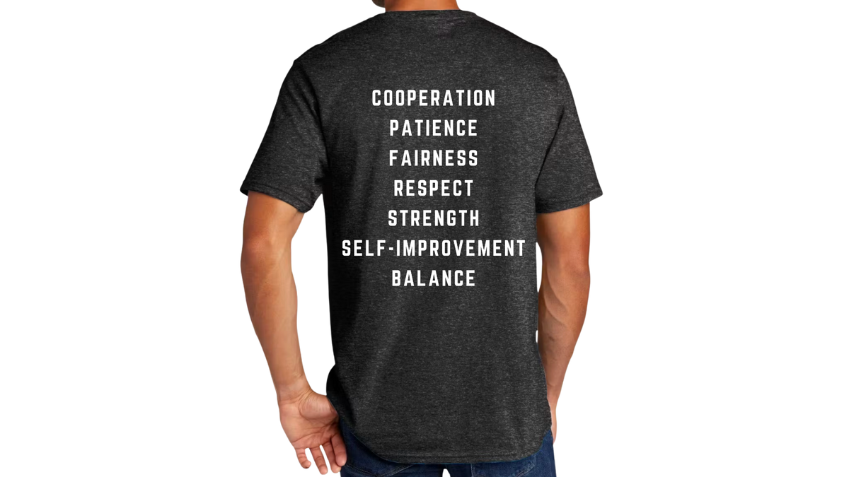 A man wearing a dark heather gray t-shirt, viewed from the back. The shirt features a vertical list of motivational words in white block lettering: 'COOPERATION', 'PATIENCE', 'FAIRNESS', 'RESPECT', 'STRENGTH', 'SELF-IMPROVEMENT', and 'BALANCE'.