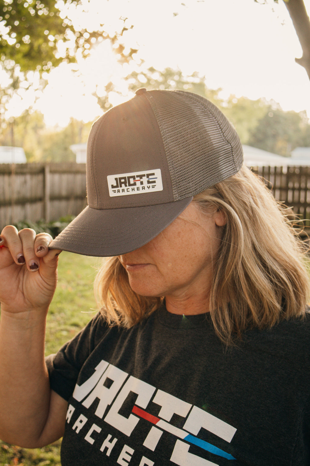 A woman outdoors wearing a dark gray JROTC Archery trucker hat and a black JROTC Archery t-shirt, holding the brim of the hat.