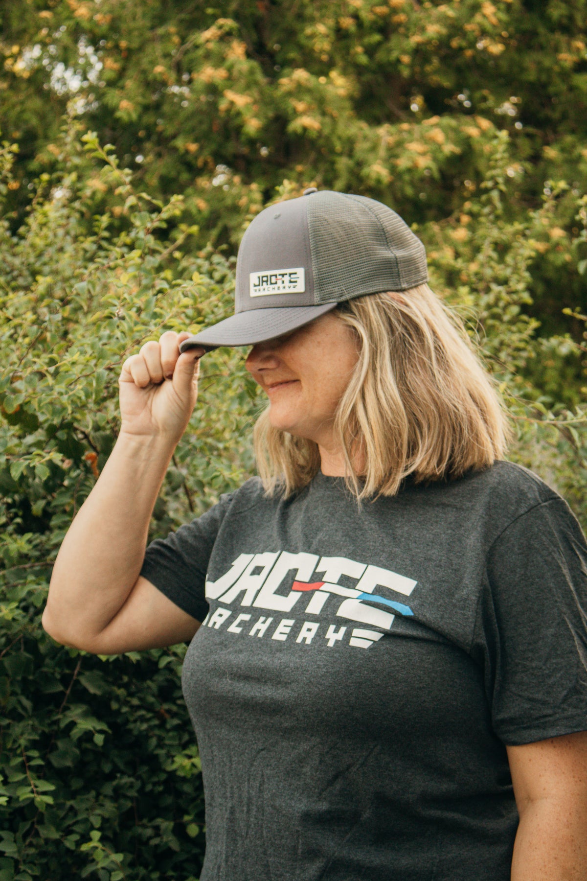A woman outdoors wearing a dark gray JROTC Archery trucker hat and a dark gray JROTC Archery t-shirt, with green foliage in the background.