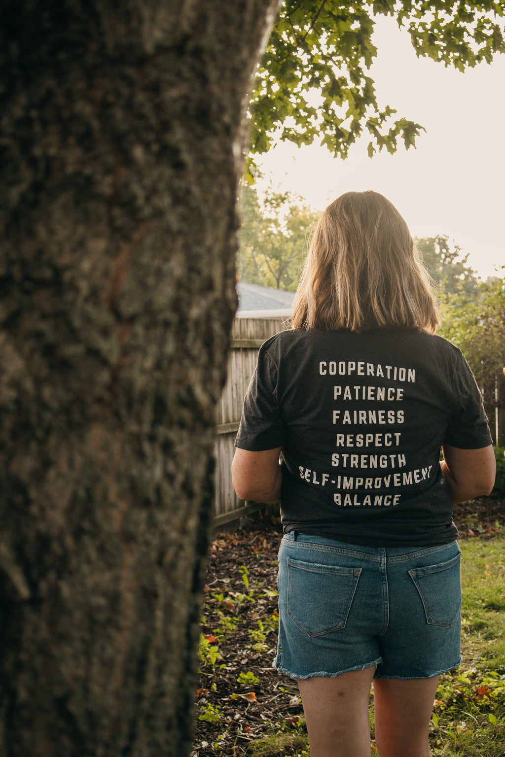 A woman viewed from behind wearing a gray t-shirt with white text listing virtues like cooperation, patience, fairness, and respect, paired with denim shorts in an outdoor garden setting.