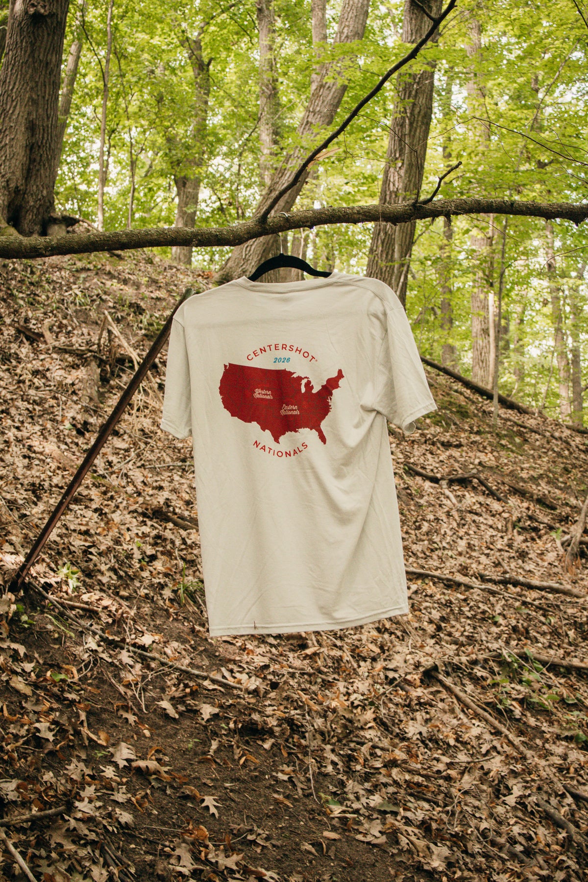 A cream-colored t-shirt hanging from a black hanger on a tree branch in a wooded forest setting. The back of the shirt features a red graphic of the United States map with the words 'CENTERSHOT 2026' and 'NATIONALS' printed in red.