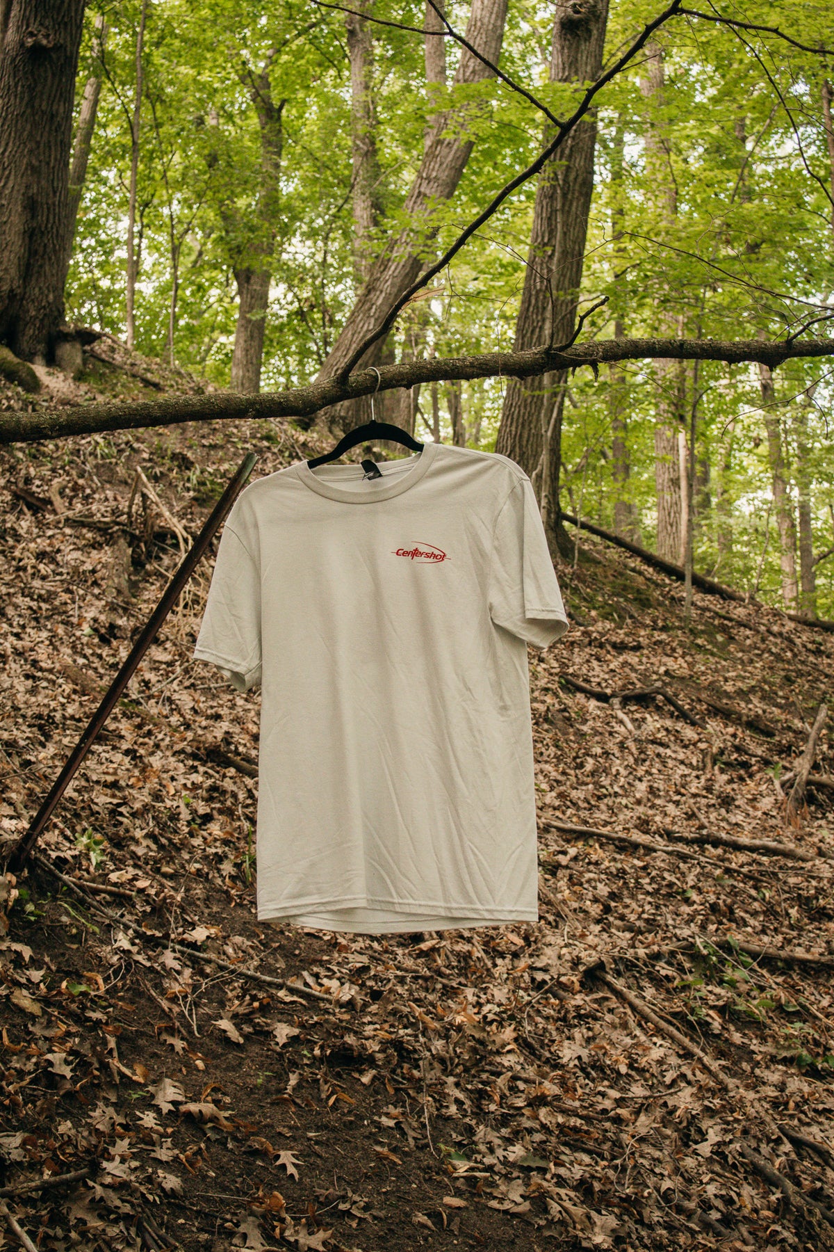A cream-colored short-sleeve t-shirt with a small red CENTERSHOT chest logo, hanging from a black hanger on a tree branch in a wooded forest setting.