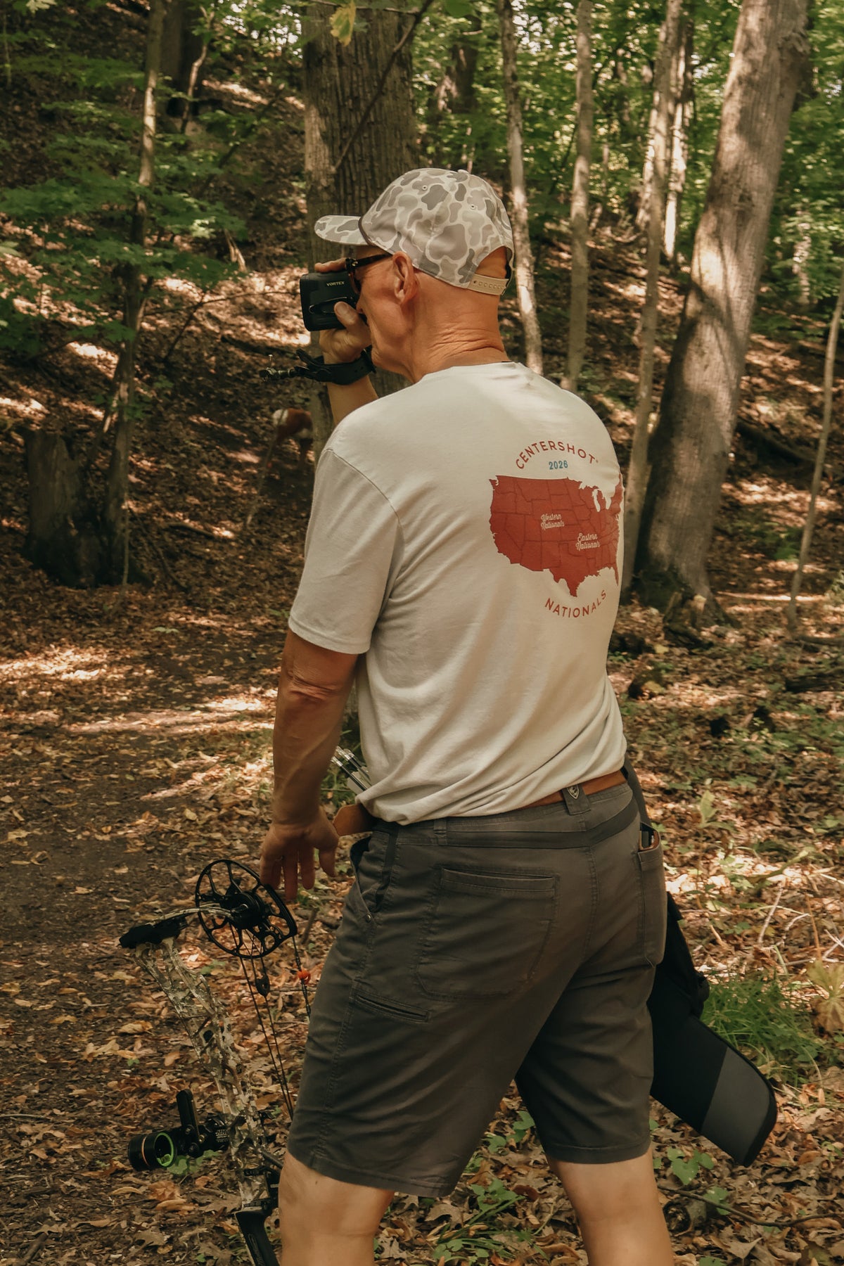 A man standing in a wooded outdoor setting, photographed from behind, wearing a light gray t-shirt with a red map graphic and the words 'CENTERSHOT NATIONALS' on the back, along with dark shorts and a camouflage baseball cap.
